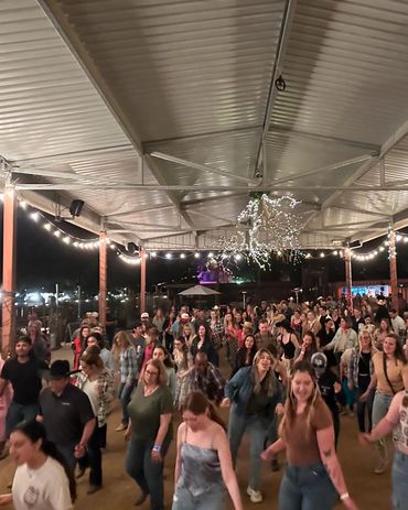 Large group line dancing under string lights in a covered outdoor area at night.