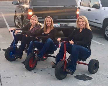 Three women happily riding small tricycles in a parking lot.