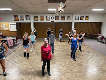 A group dance class practicing in a community hall with portraits on the wall.