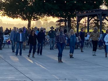Group of people line dancing outdoors at sunset with string lights above.