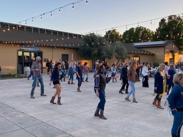 People line dancing outdoors near a building with string lights at dusk.