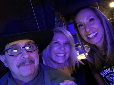 Three people smiling under blue light, one man wearing a black hat and glasses.
