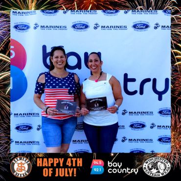 Two women smiling with certificates, celebrating 4th of July at Bay Country event.