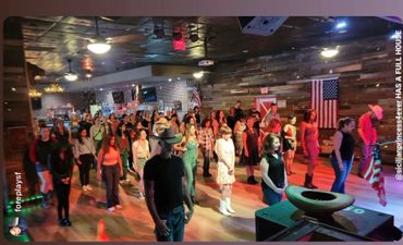 Group of people line dancing indoors with American flags and cowboy hats.