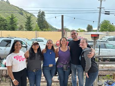 Group of six friends smiling outdoors with mountains in the background.