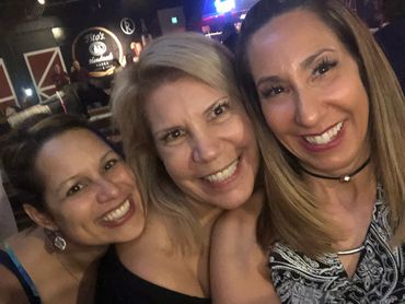 Three women smiling happily together in a lively bar setting.