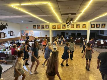 Group line dancing in a decorated community hall with portraits on the wall.
