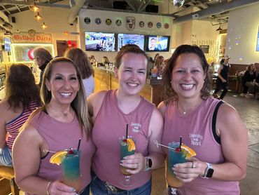 Three women in matching pink tops holding colorful cocktails and smiling indoors.