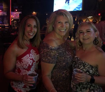 Three women smiling at a dimly lit bar, holding drinks and enjoying the night.