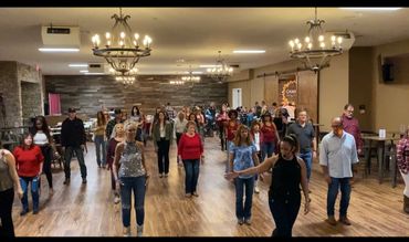 A large group learns line dancing in a spacious, warmly lit room.