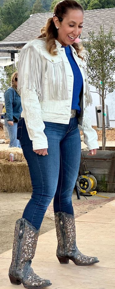 Woman smiling in a fringed jacket and decorative cowboy boots outdoors.