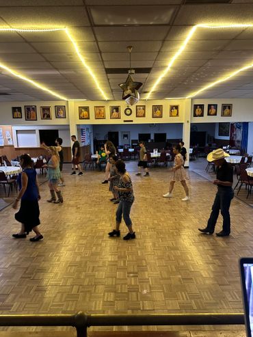 People line dancing in a community hall with wooden floors and framed portraits.