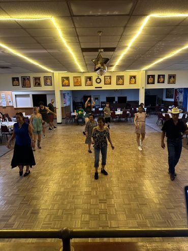 Group of people lined up practicing dance steps in a hall with portraits on the wall.