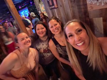 Four women smiling and posing for a selfie at a lively indoor event.