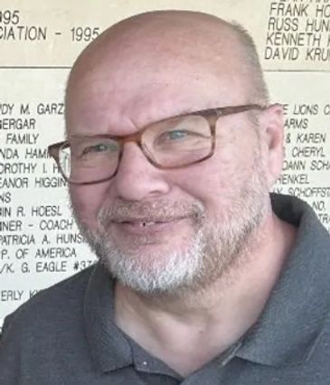 Smiling middle-aged man with glasses and beard in front of a text-covered background.