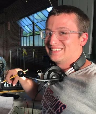 A smiling man with glasses and headphones around his neck holding a pencil.