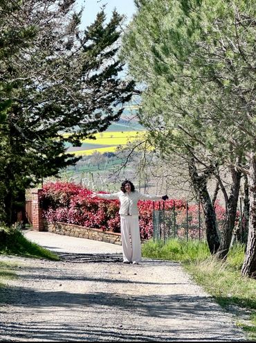 A woman stands on a sunlit path with arms outstretched, surrounded by trees and colorful foliage.
