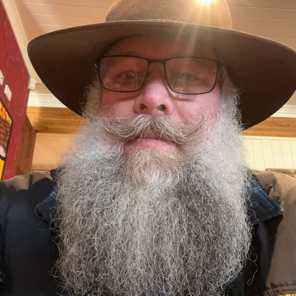 Man with a long gray beard and glasses wearing a brown hat.