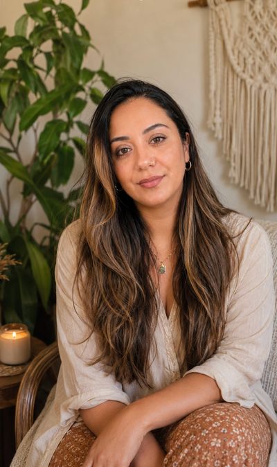 A woman with long hair sitting indoors with a plant and a candle in the background.