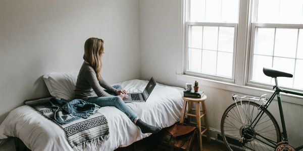 A woman sitting on a bed working on a laptop near a window with a bicycle beside.