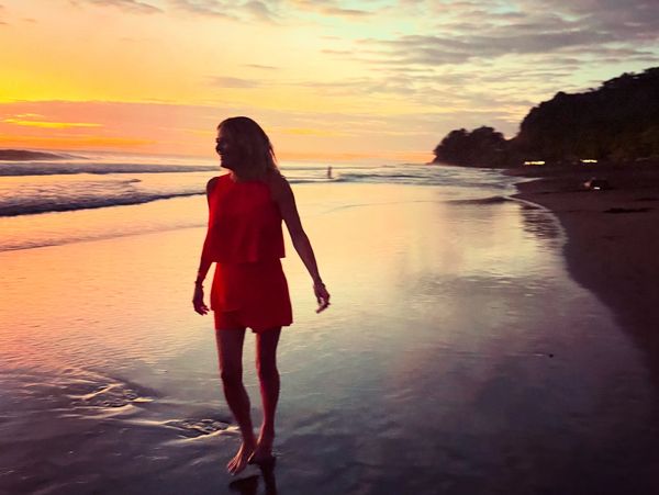 Woman in red dress walking barefoot on a beach at sunset.