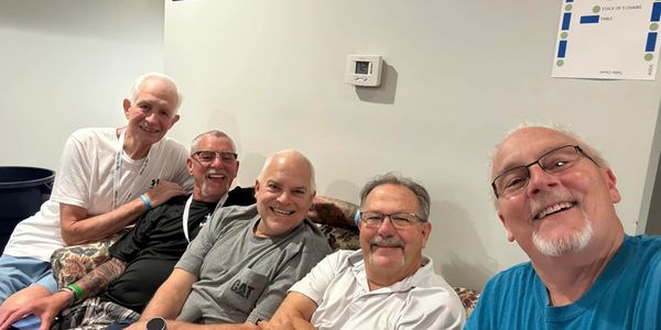 Five older men smiling and sitting closely on a couch indoors.