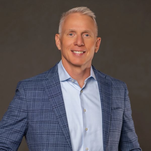 Smiling man in a blue plaid blazer and light blue shirt against a plain background.