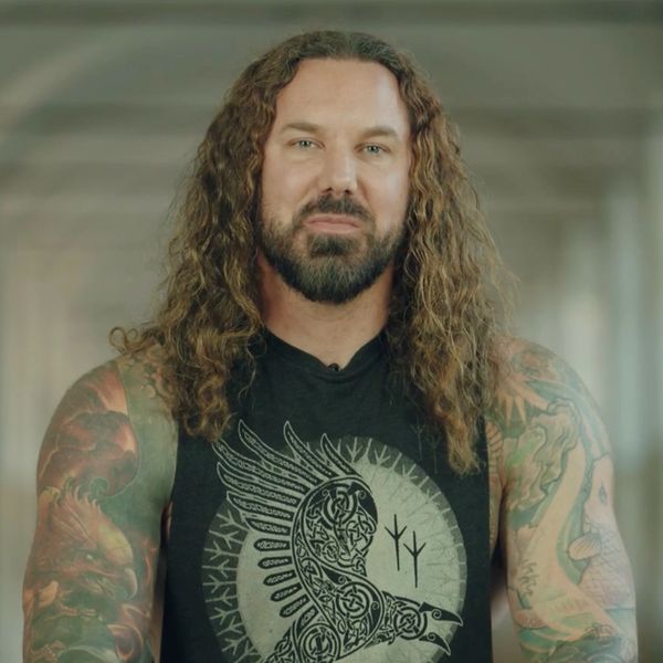 Man with long curly hair and tattoos wearing a sleeveless black shirt with a raven design.