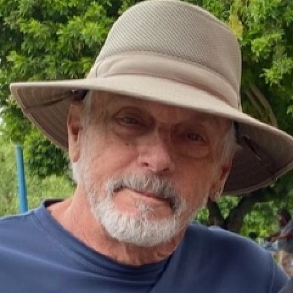 Elderly man with a white beard wearing a wide-brimmed hat outdoors.