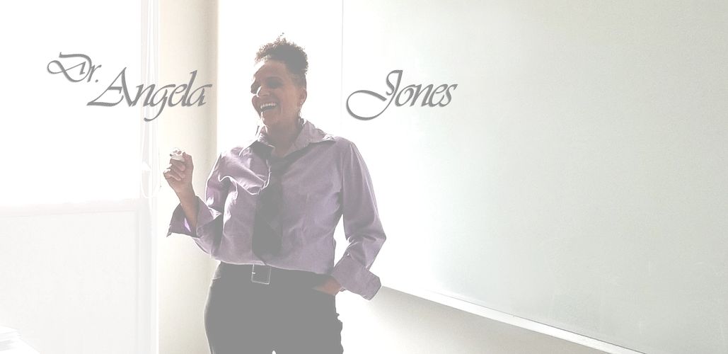 Jones is standing in front of a whiteboard in a classroom. She is wearing a purple shirt and a tie.