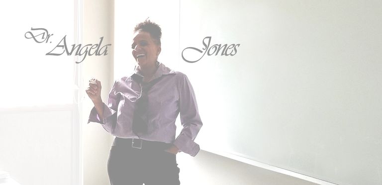 Jones is standing in front of a whiteboard in a classroom. She is wearing a purple shirt and a tie.