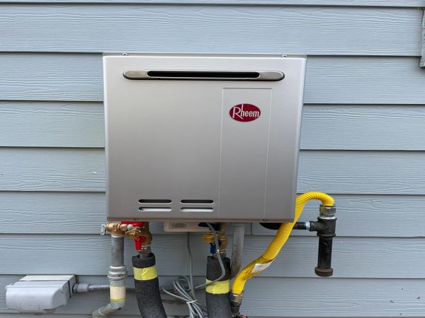 A Rheem tankless water heater installed on a gray wooden exterior wall.
