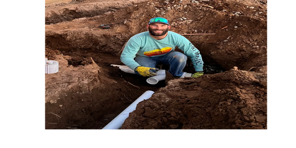 A man working on a plumbing pipe in a dirt trench outdoors.