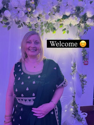 A smiling woman in traditional attire stands under floral decorations with a welcome sign.