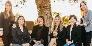 A group of six women posing outdoors in professional attire.