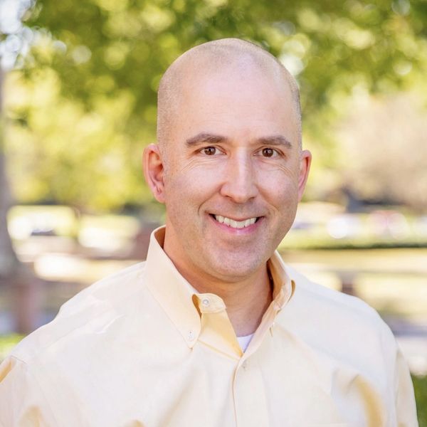 Smiling bald man in a light yellow shirt outdoors with greenery in the background.