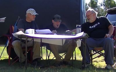 Three men sitting outdoors around a table with papers, engaged in discussion.