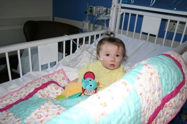 Baby in hospital bed with nasal oxygen tube covered in pink and teal quilt.