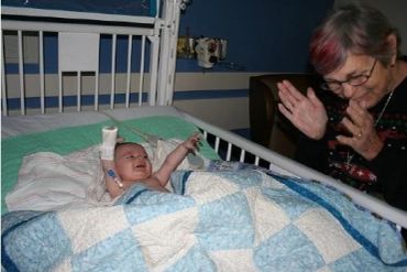 Grandmother playing with baby grandson who is lying in a hospital crib covered with a blue and white quilt.