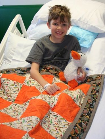 Smiling boy in hospital bed covered with an orange and brown quilt.