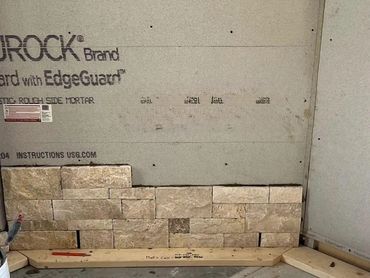 Partially installed stone brick wall on cement board in a construction area.