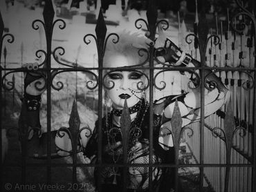 A pale woman in 80s deathrock attire poses behind a cemetery fence gate