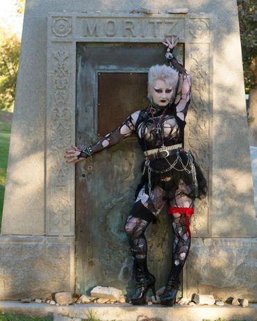 A pale woman in 80s deathrock attire poses in front of a mausoleum 
