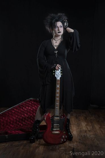 A pale woman in 90s goth attire poses with a coffin shaped guitar case and red SG guitar