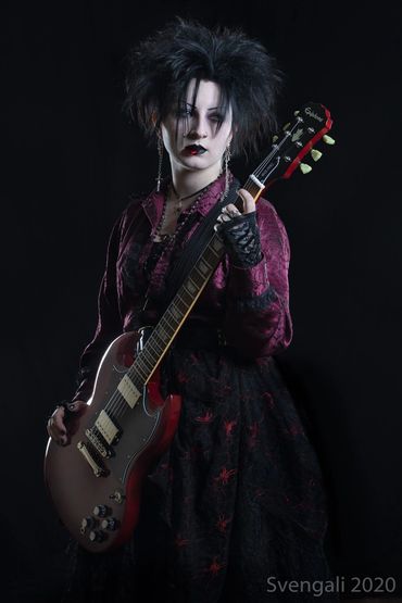 A pale woman in 90s goth attire holds a red guitar in her hands and a guitar pick in her mouth