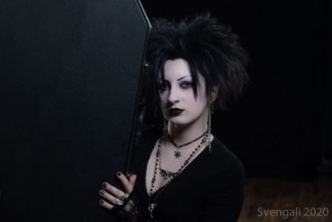 A pale woman in 90s goth attire poses with a coffin shaped guitar case