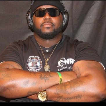 Muscular man with sunglasses and headphones, arms crossed, showing tattoos and wearing a watch.