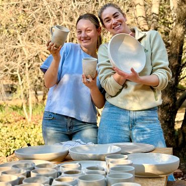 Two women holding handmade pottery with many ceramic pieces on the table.