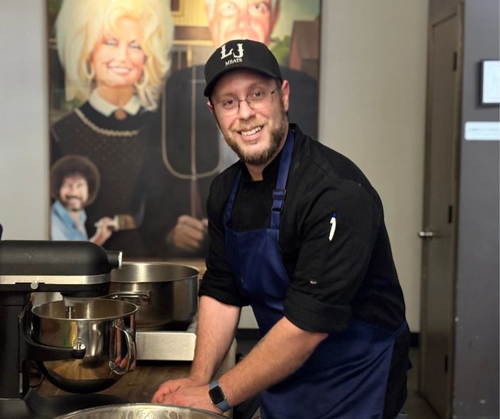 Smiling chef in a blue apron and working in a kitchen in front of Anthony Bourdain and Dolly.