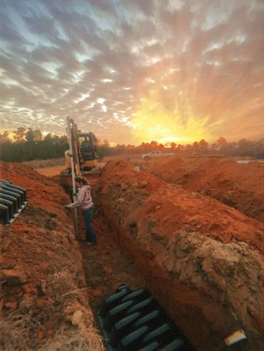 Person working in a deep trench during a vibrant sunset with construction equipment.